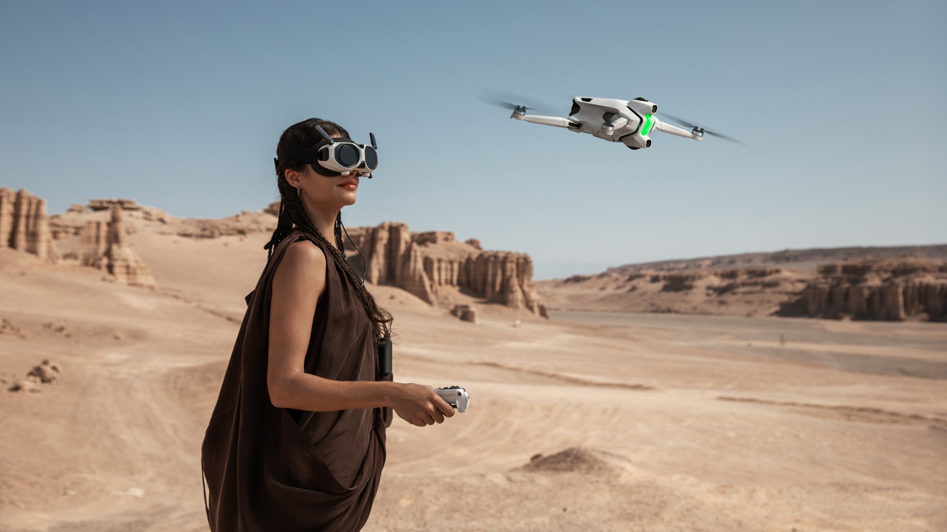 A woman wearing a brown dress and goggles holds a remote and looks up at a white drone with green lights flying in a vast, dry canyon.