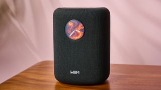 The Wiim Sound speakers shown from the front, sitting on a wooden table. It has a small circular screen on the front near the top, which shows a clock on a firey background.