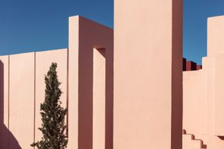 Close up view of the pink exterior walls at La Muralla Roja by Ricardo Bofill in Calp, Spain. There is a tree outside the building and the sky is clear and blue