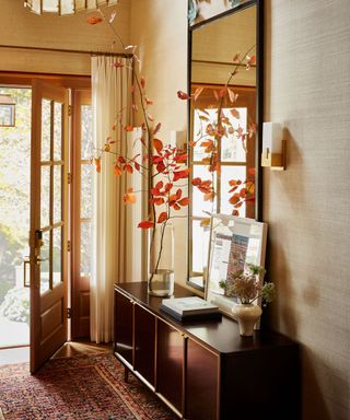 entryway with brown side cabinet, large mirror, neutral walls and autumnal foliage