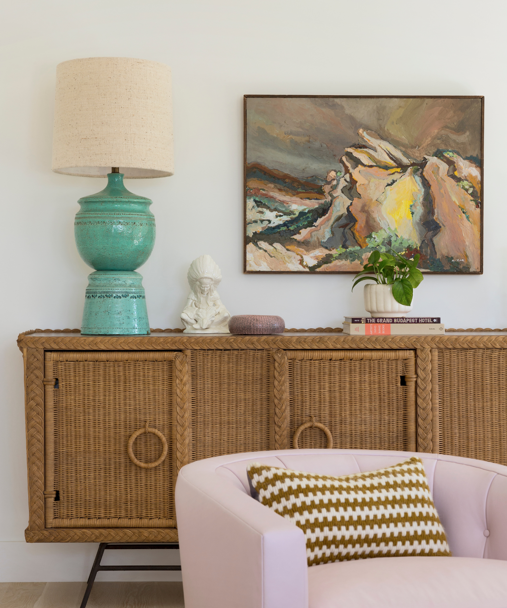 a rattan credenza holding a turqoise lamp, some decorative objects, and a small plant. In front of the credenza is a light pink chair with a brown and white pillow