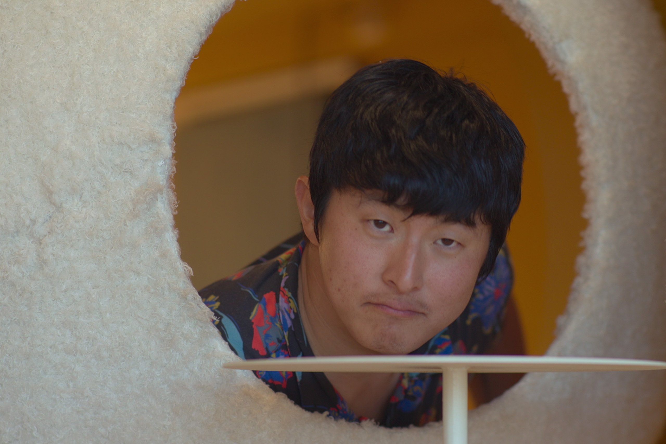 A man (Korean TV personality Kian84) has a neutral expression as he peeks through a circle cutout on a white, fluffy wall. A small white bench is in front of him and the background is yellow behind him. A TV still from 'Kian's Bizarre B&amp;amp;B' season 2.