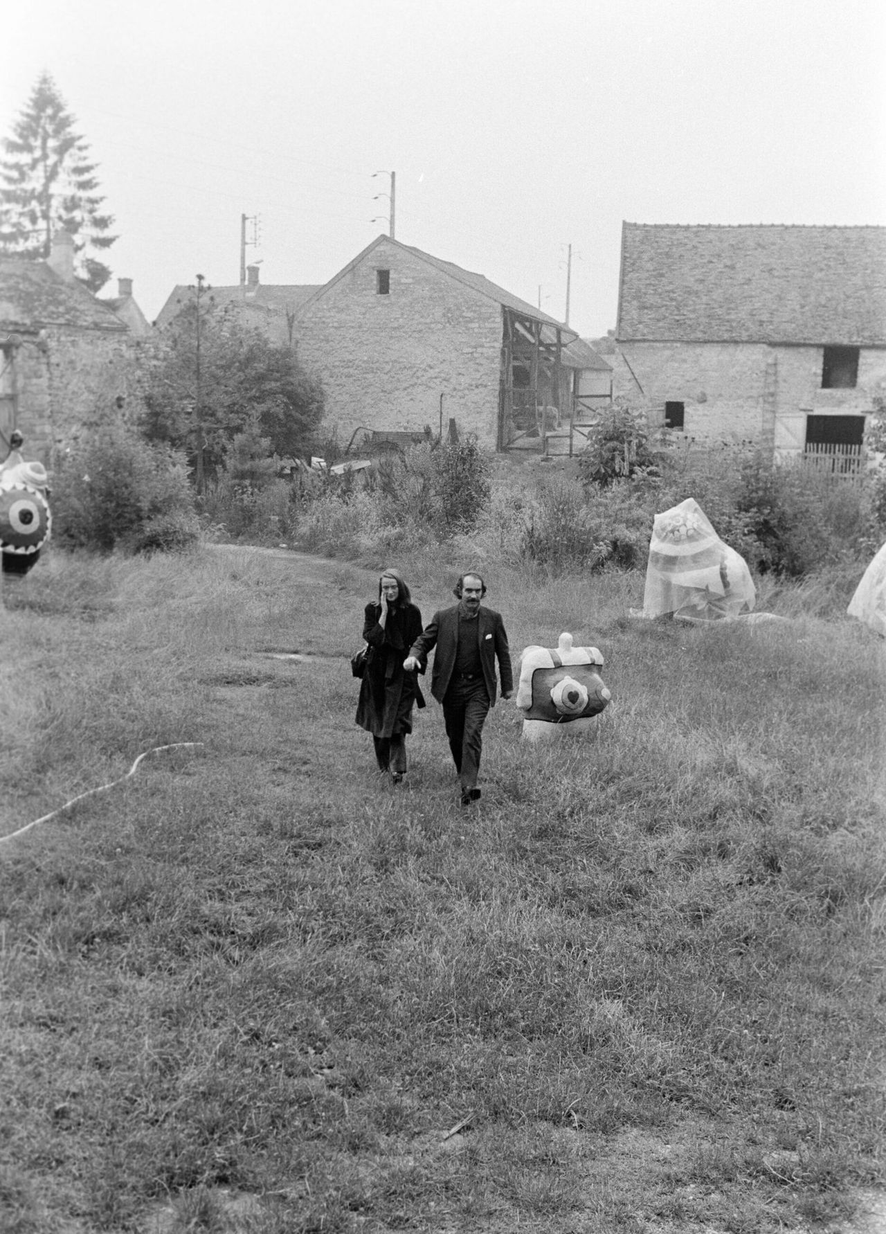 A woman and a man walk in a field filled with whimsical, creature-sque sculptures in a black and white photograph.