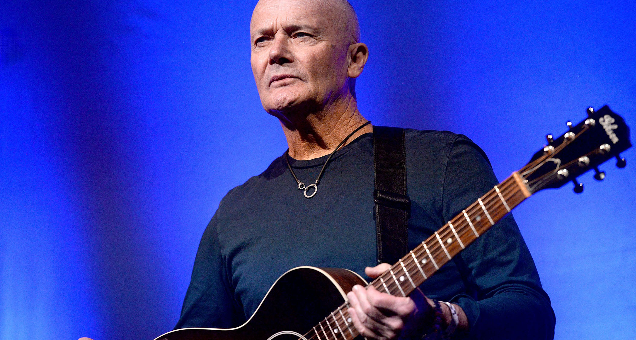 Creed Bratton plays a Gibson acoustic onstage against a deep blue background.