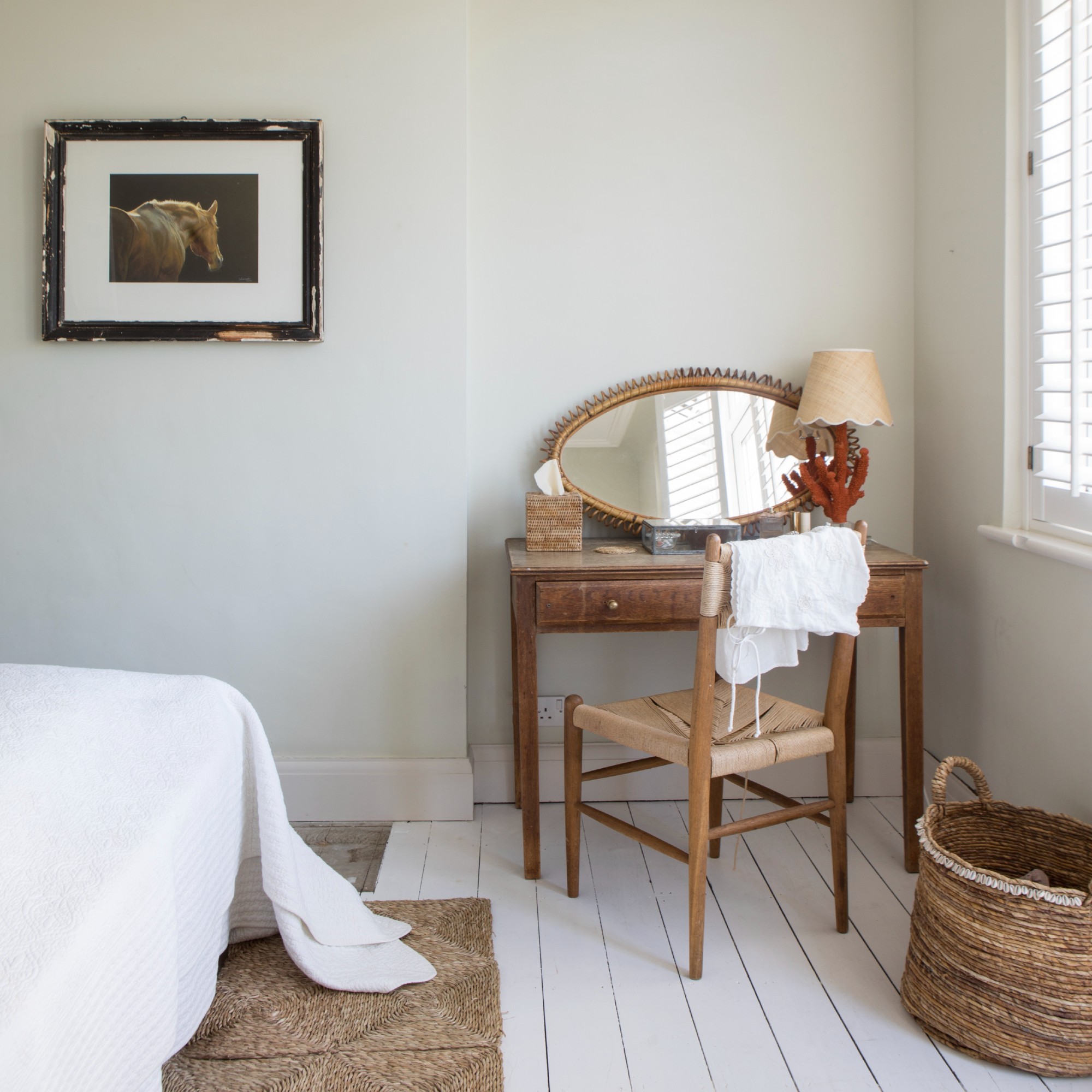 A light bedroom furnished with vintage furniture, including a dressing table with an oval mirror and a table lamp by the window
