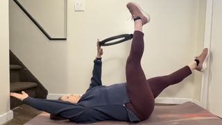 Jennifer Rizzuto performs deadbug with a Pilates ring. She is lying with her back on the floor. Her left hand and right leg are pointing up and she holds a Pilates ring between them. Her right arm and left leg are lowered just above the ground.