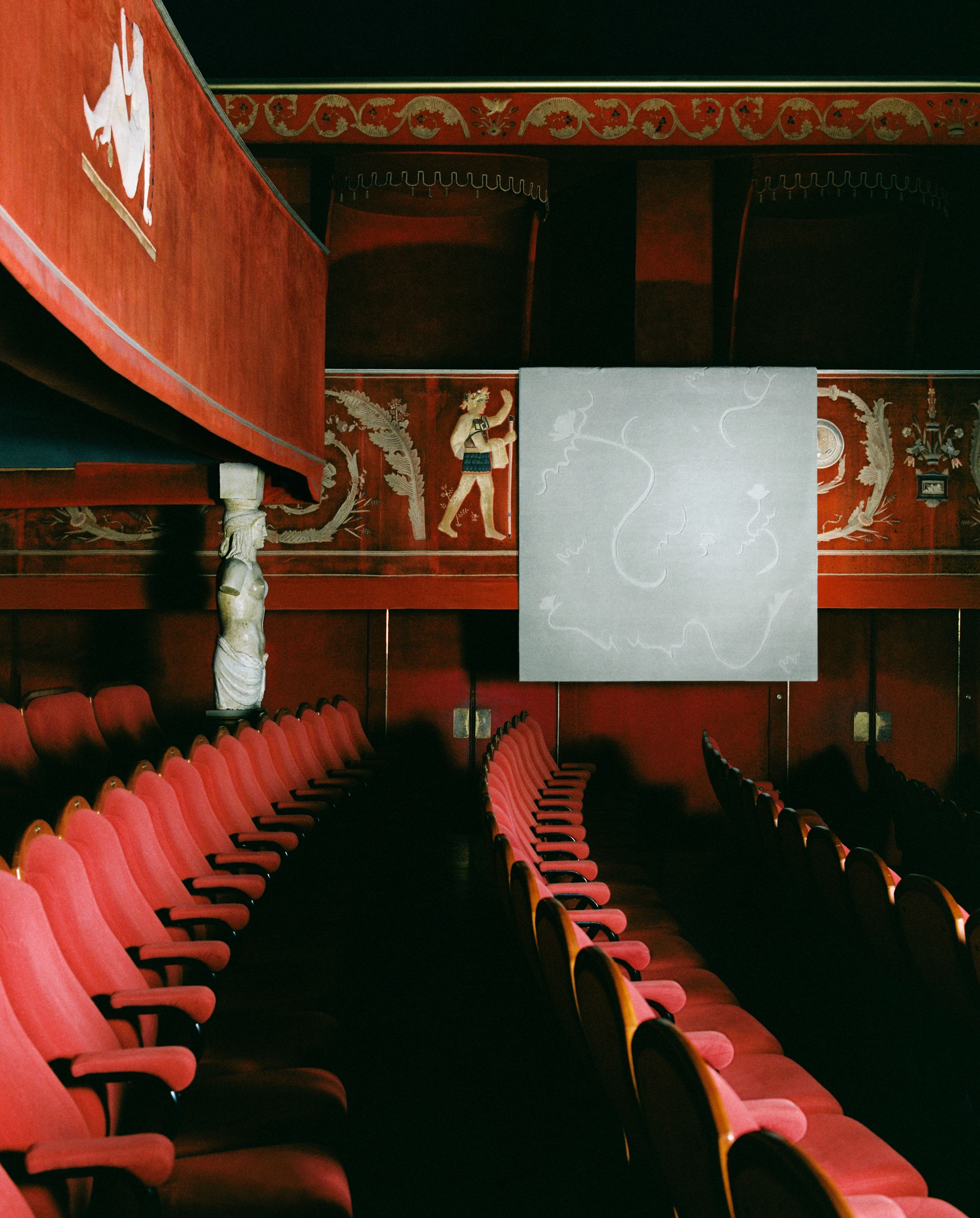 Image of a red theater with rows of seats and balconies with heritage embroidery. There is a light blue rug hanging over part of the balcony.