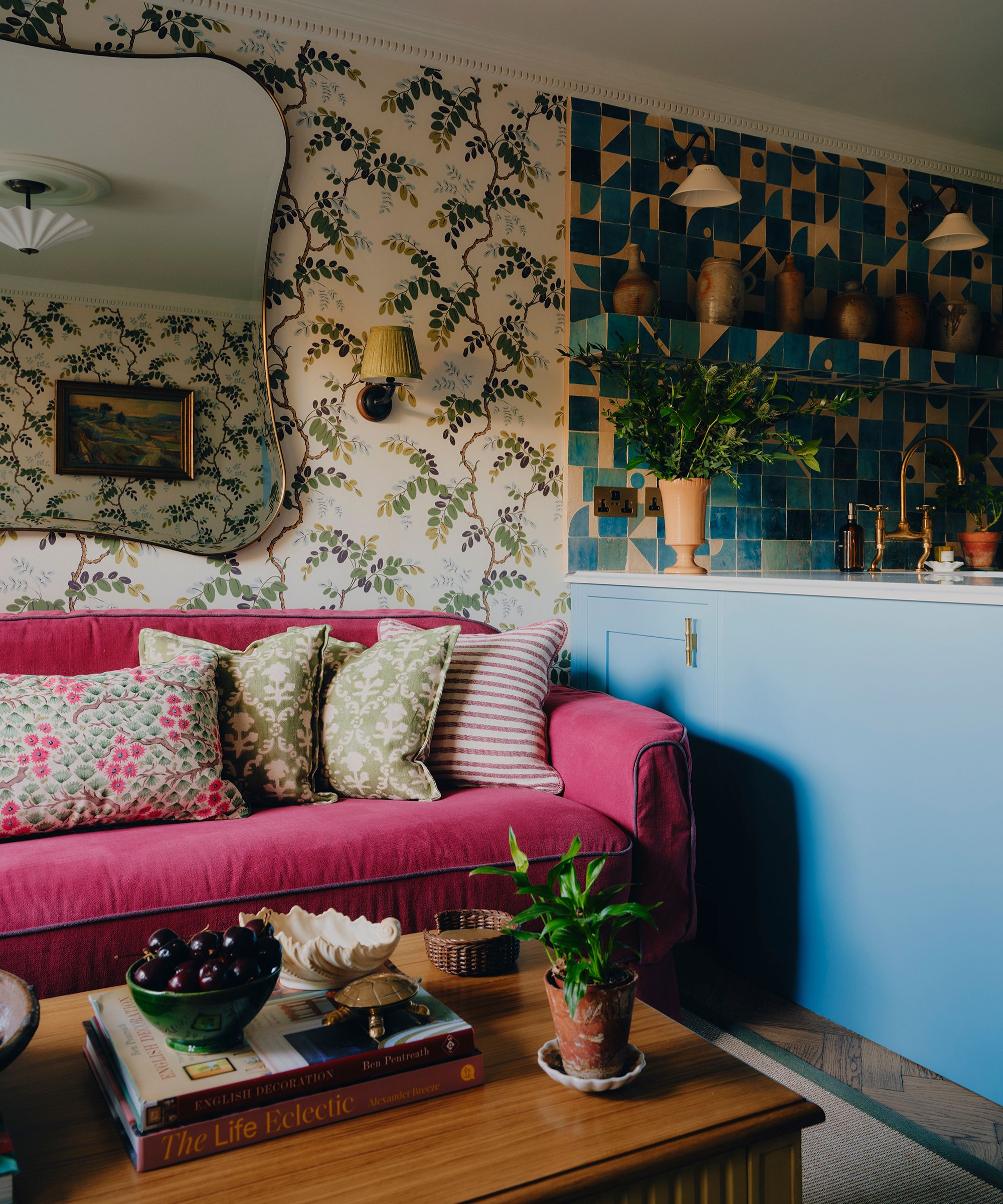 an eclectic, small living room in london with patterned wallpaper and a large dark pink couch with patterned pillows
