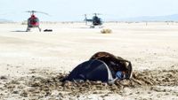 A small pile of metal parts is half buried in a sandy beach as helicopters approach from the background