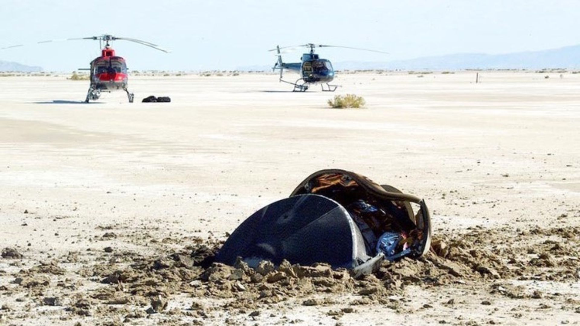 A small pile of metal parts is half buried in a sandy beach as helicopters approach from the background