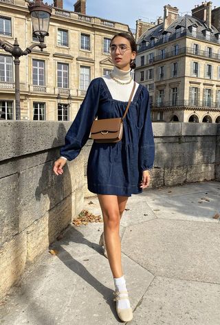 A woman's jean dress with a puff-sleeve mini layered over a white turtleneck and styled with a gold chain necklace, brown crossbody bag, and white Mary Janes.