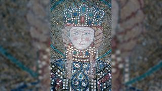 A mosaic of Empress Irene of Athens. She has blue eyes, rosy cheeks, and long blonde hair in two plaits. She is adorned with a large golden crown with blue and green jewels, with a large red jewel in the center, as well as red teardrop earrings, all on a gold background. She is wearing an intricately decorated gown.