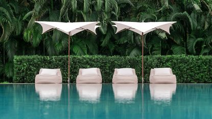 Two white patio umbrellas shading four sun loungers by the side of a turquoise blue swimming pool with lush green tropical foliage in the background 