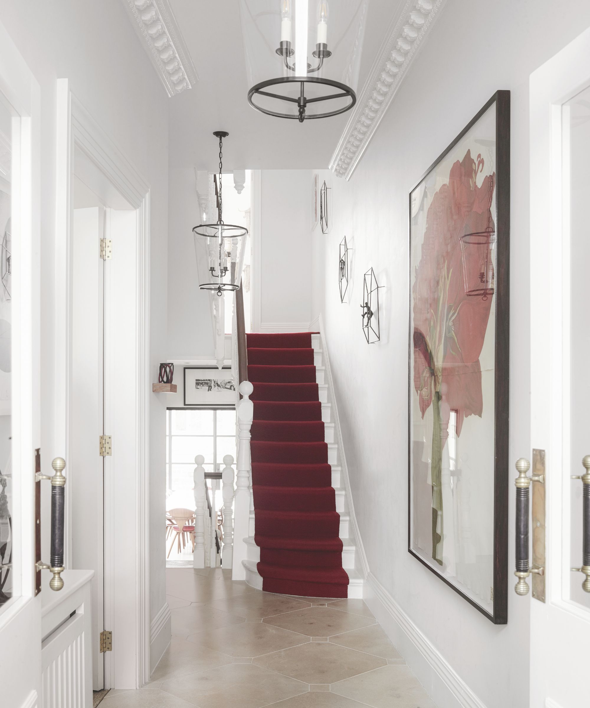 A white entryway with a white staircase with red stair runner, a large framed artwork on the wall, and glass lamps.