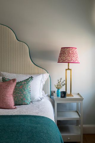 Bedroom with turquoise throw, upholstered headboard with turquoise edging, gray painted side table, lamp with patterned shade, red and turquoise cushions, white walls