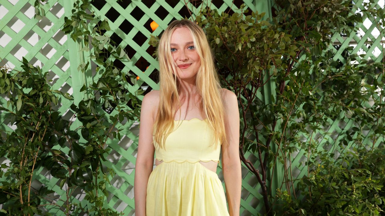 Dakota Fanning with her hair down wearing a yellow strapless dress, standing in front of a green screen with plants around it