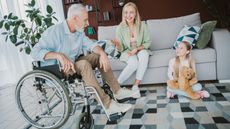 An older man in a wheelchair, his adult daughter and her young daughter smile as they talk in the living room.