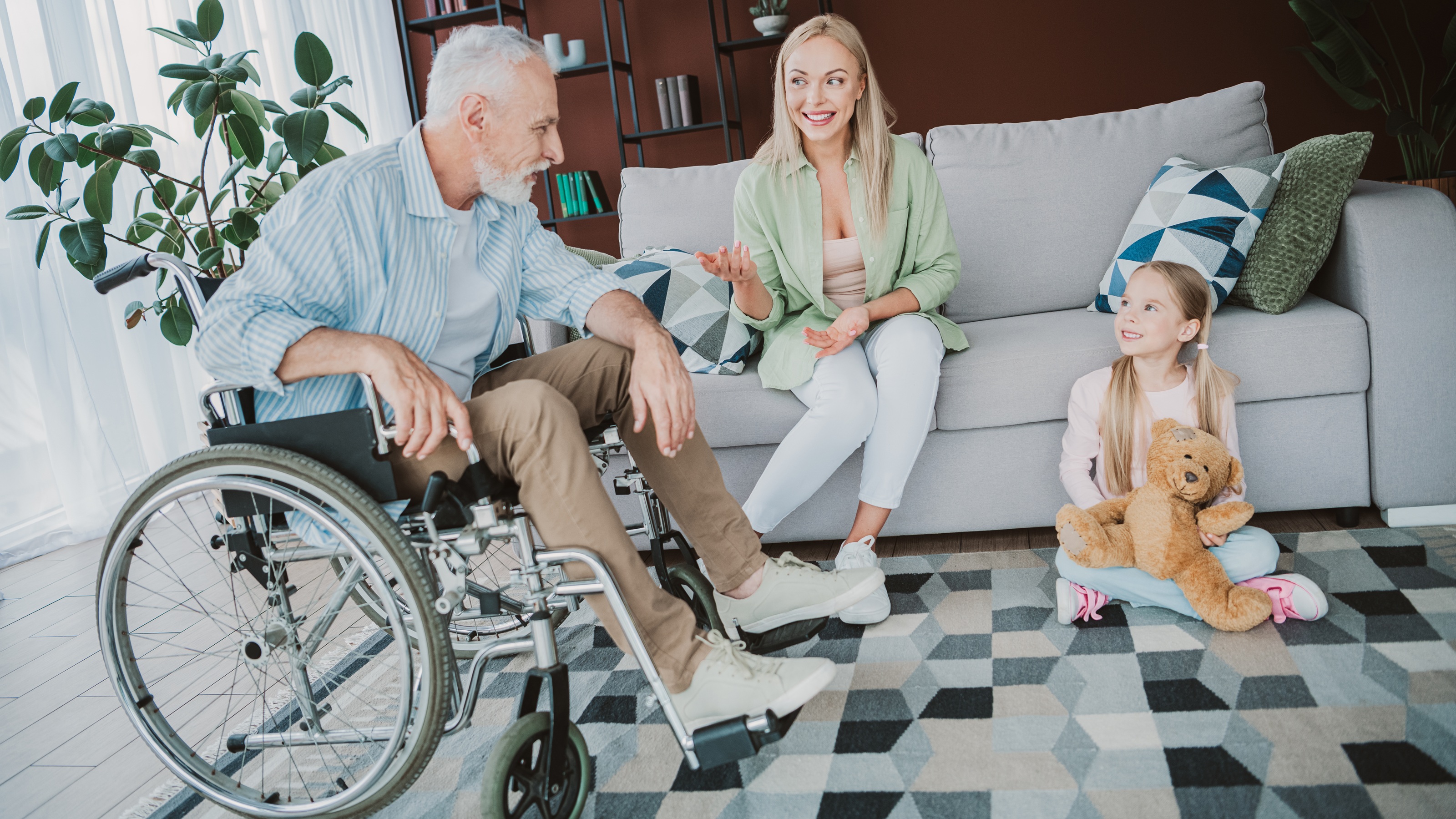 An older man in a wheelchair, his adult daughter and her young daughter smile as they talk in the living room.