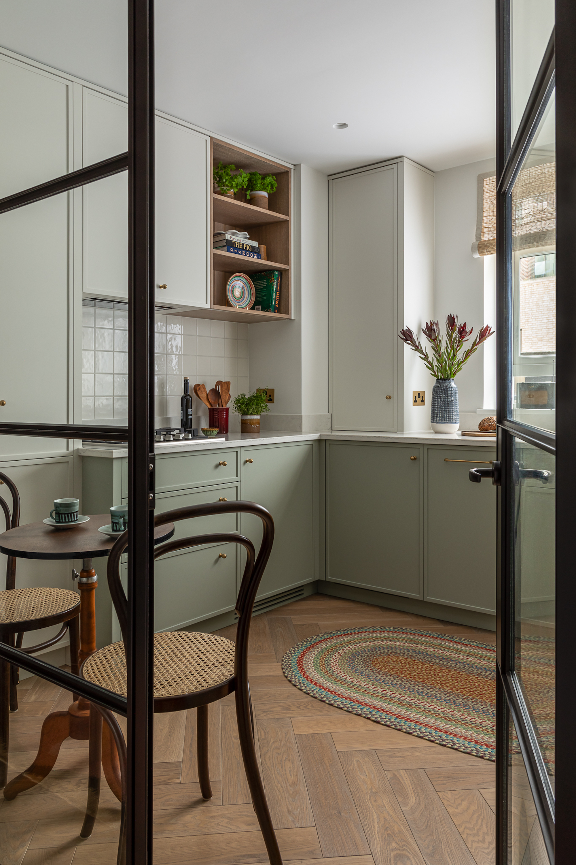A pale olive colored kitchen with light wood, herringbone flooring. There is a glass divider and door with black detailing, leading through, with a small, wooden table to the left.