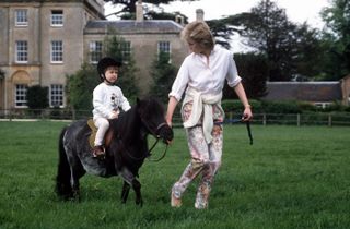 Princess Diana leading Prince William, wearing a Mickey sweatshirt and black helmet, on a brown pony through grass