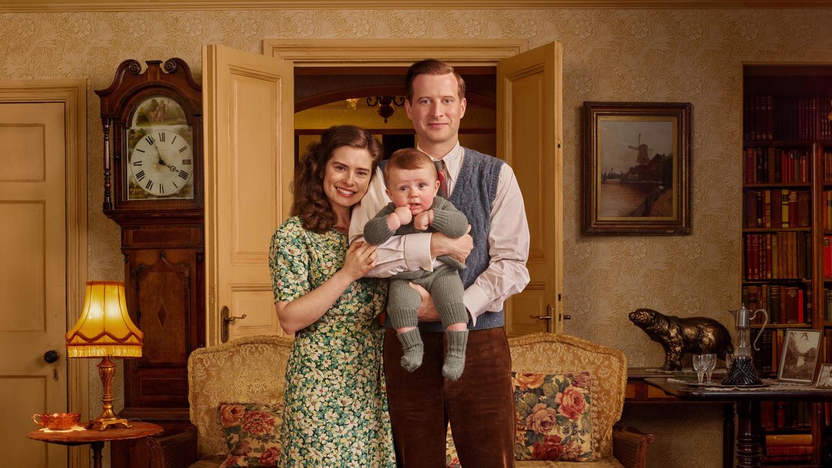 Rachel Shenton in a green floral dress as Helen stands with Nicholas Ralph in a grey tank top holding a baby in Skeldale House in All Creatures Great and Small