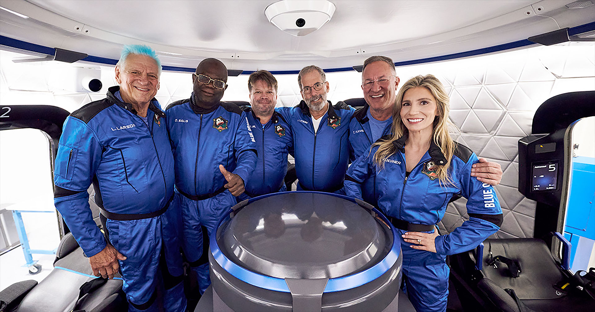 five men and a woman, all dressed in blue and black flight suits, stand inside a mockup of a space capsule