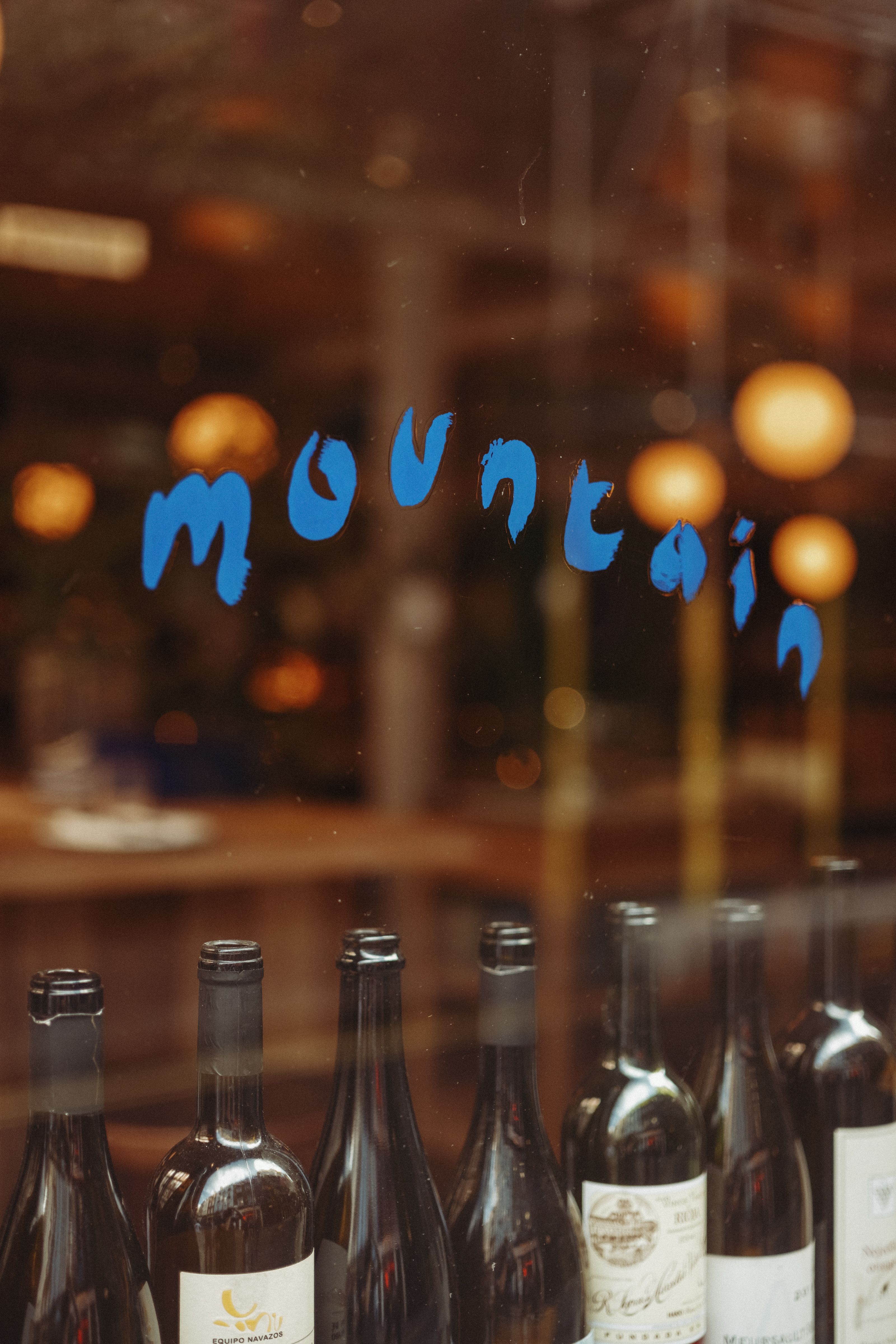 A series of wine battle are positioned behind a glass facade of a restaurant that bears a hand-drawn blue sign that reads "mountain".