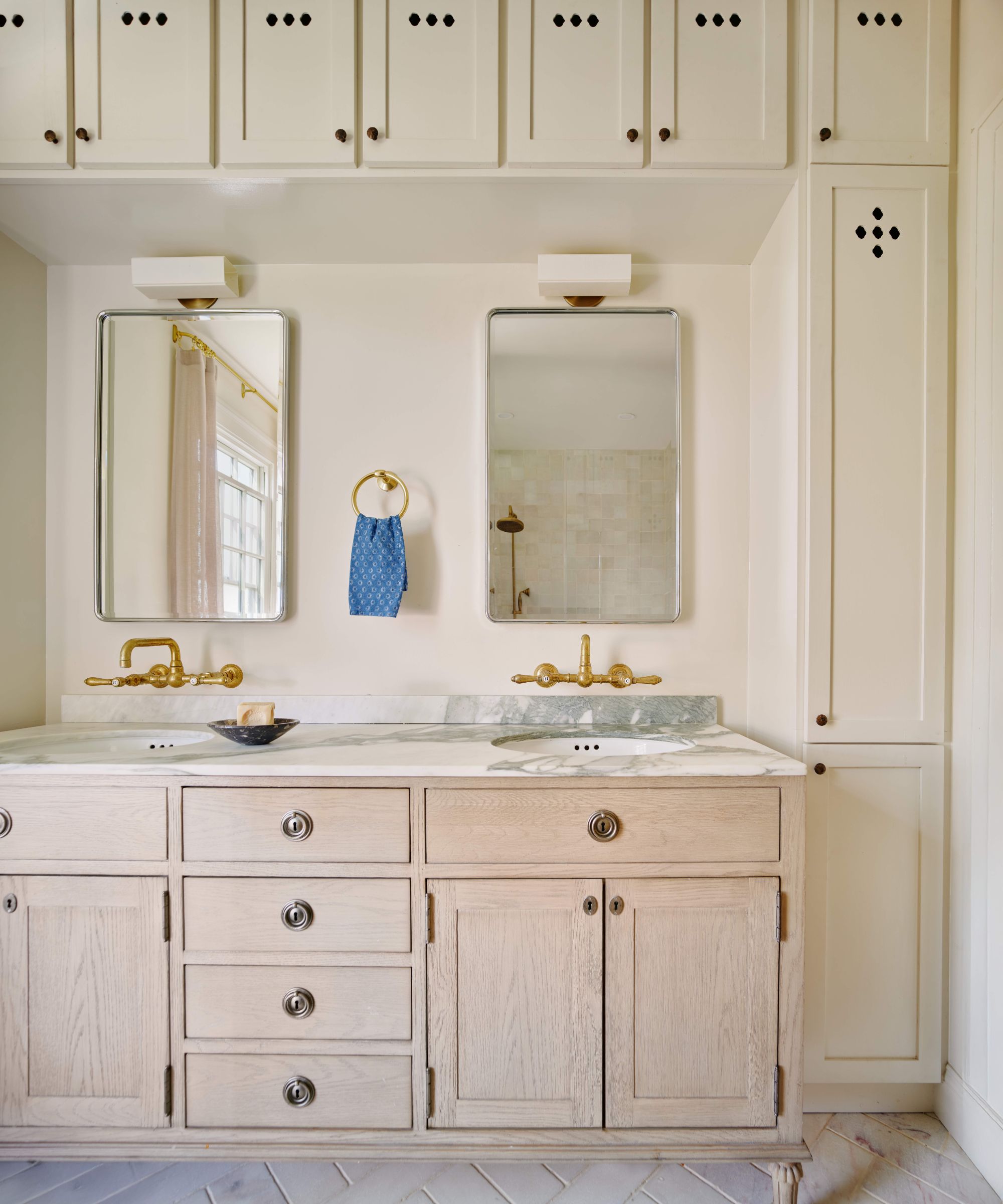 Bright double vanity bathroom with light wood cabinets, marble countertop, brass faucets, rectangular mirrors, and floor-to-ceiling storage cabinets with small diamond cutouts
