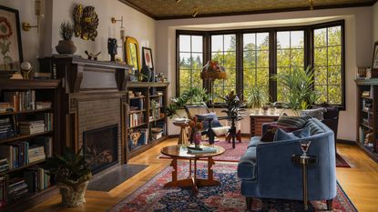 a large home library with original tudor bay window, a large vintage rug, blue sofa, accent wallpapered ceiling and wooden fireplace with bookcase built-ins
