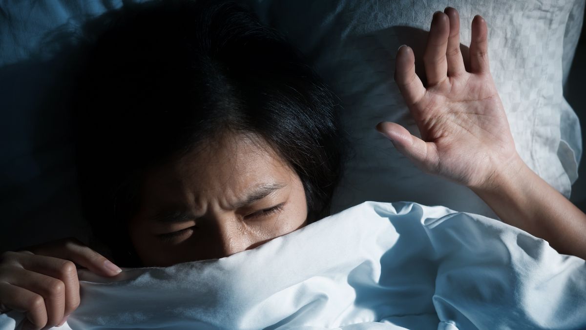 photo of a young woman lying in bed and covering her face with a blanket, as if having woken up from a nightmare