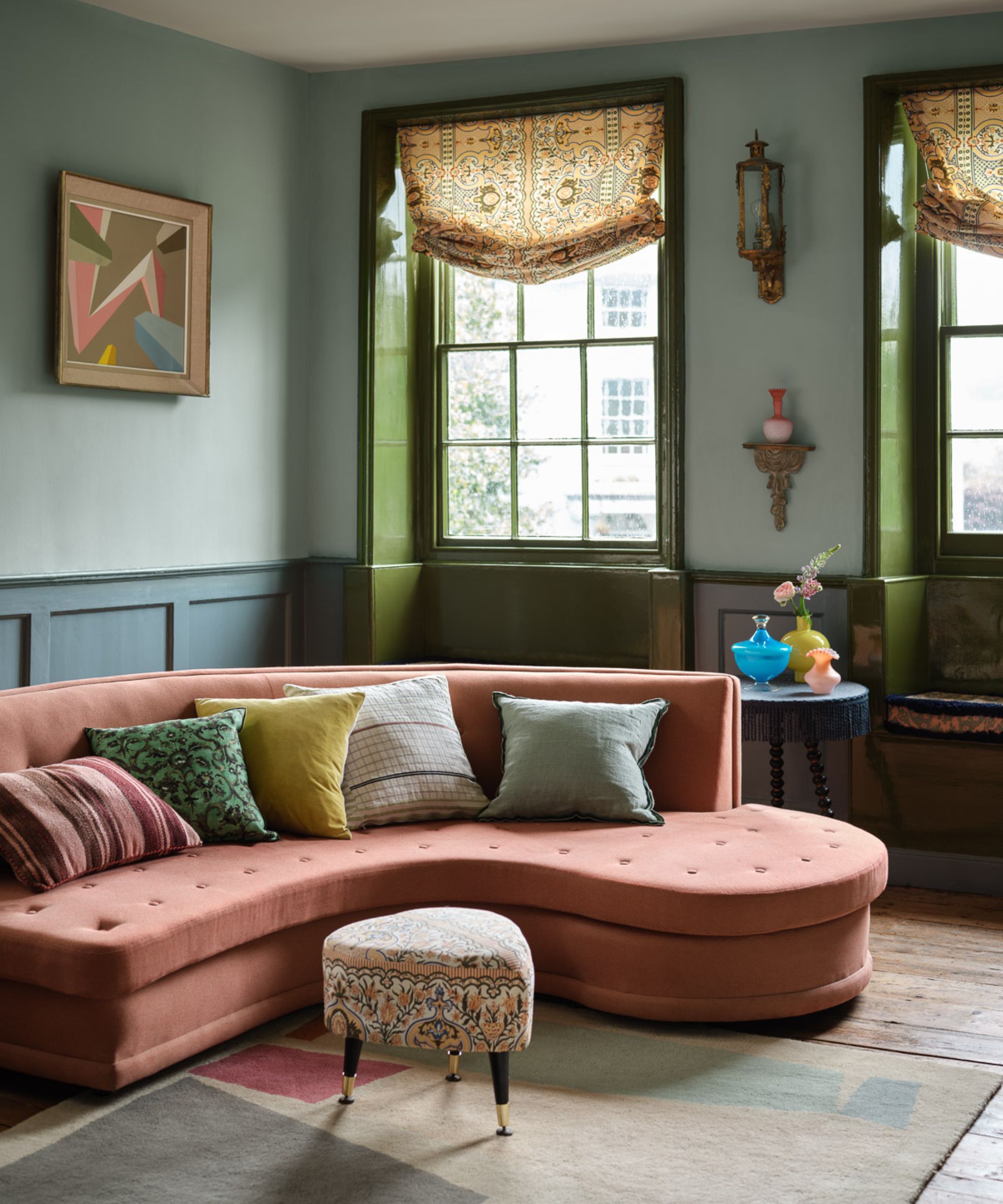 Blue painted living room with curved sofa and vintage accents including heart shaped foot stool and patterned window treatments.