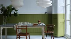 Image of a dining room with a rustic wooden table painted white with a blue worktop. There are two wooden chairs around the table and two white paper lanterns hanging above it. The wall and window shutter are painted light blue and olive green in a color-blocked fashion.