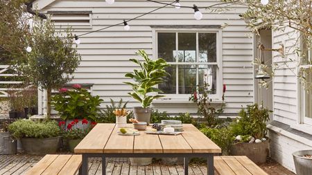 Image of the exterior of a home made of wood panels that are painted white. There are cafe lights hung around a patio with a wooden dining table and matching bench seats. There are lots of plant pots.