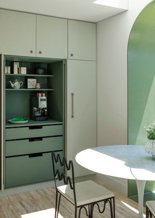 A bright green kitchen with hidden pantry storage that opens up. In the kitchen is also a bright white dining table and a curved black metal chair with a white cushion seat.