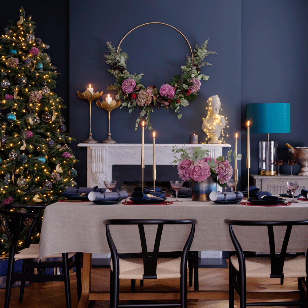 gold decorative hoop above a mantel in dark blue dining room behind set table with tall candles next to Christmas tree