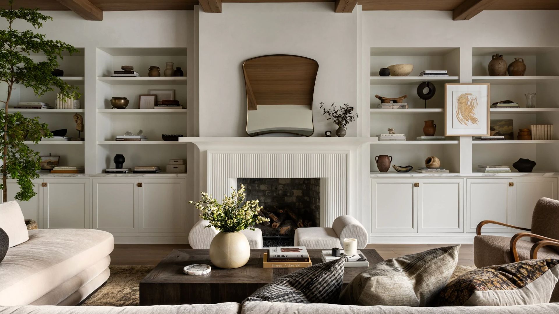 A white living room with built-in shelves on either side of a fireplace, featuring ceramics, bowls, books, and candles, in front of a coffee table with a vase of flowers around a beige sectional, a pair of armchairs and cushions 
