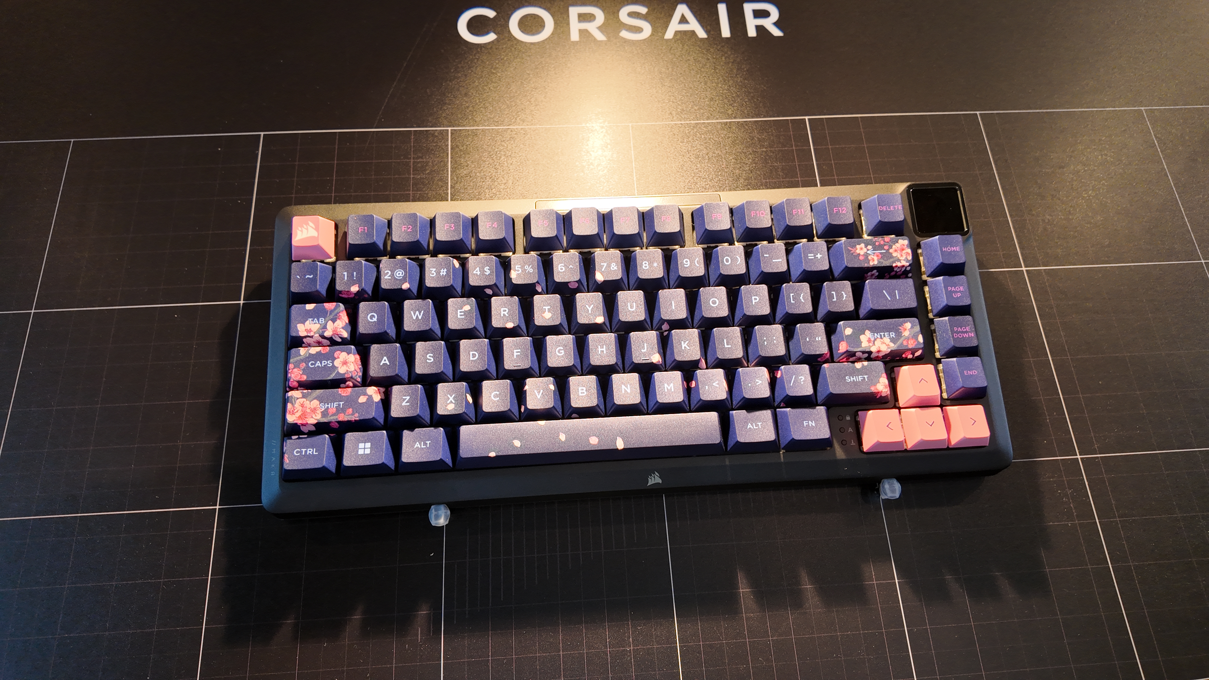Corsair's MAKR 75 keyboard as seen on the show floor at Computex 2025. The keycaps show the 'dark cherry' design, with purple keycaps accented by a pink cherry blossom motif. This keyboard has the LCD screen add-on module in the top right-hand corner.