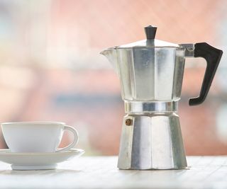 A stainless steel moka pot on a countertop by a cup