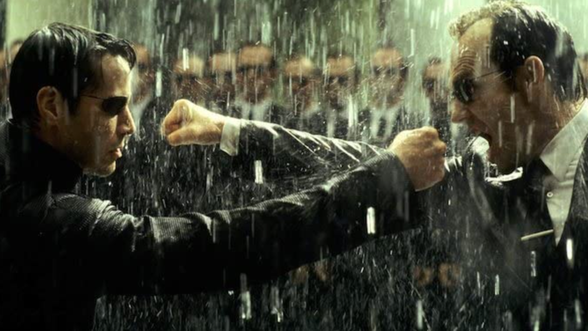 A still from The Matrix Revolutions movie showing two men fighting in the rain. On the left we see Neo, wearing a black trench coat and sunglasses with this right arm outstretched in a punch. On the right is Agent Smith, wearing a suit and sunglasses, with his right arm outstretched in a punch. In the background you can see an army of Agent Smiths.