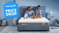 The Tempur-Pedic Tempur-Cloud mattress on a bed frame in a bedroom, a happy couple sits on top of the bed, a Tom's Guide price drop deal's graphic (left)