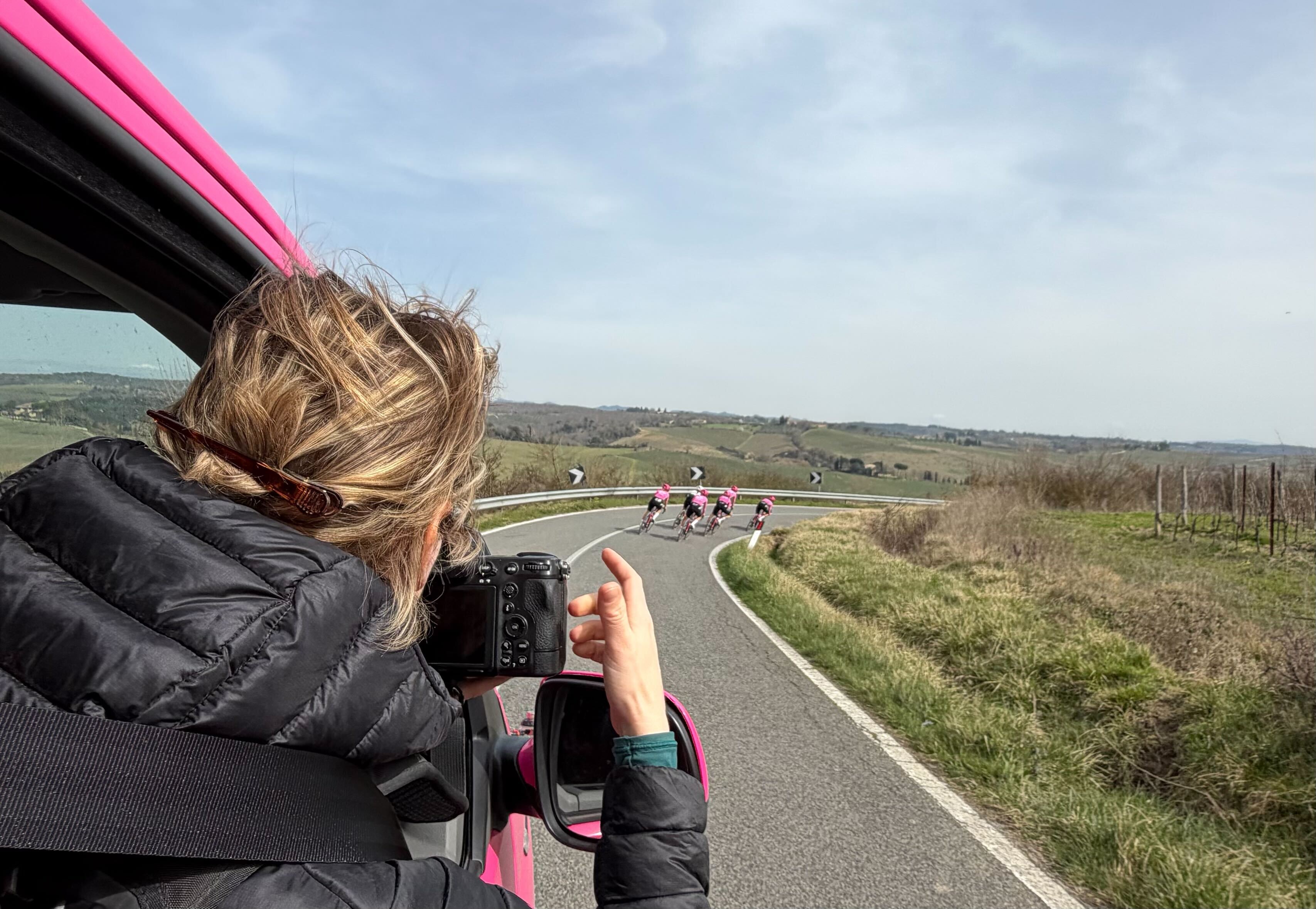 A woman leans out of the team car to take a photo of the EF riders descending in the distance