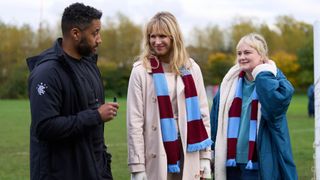 Samuel Anderson in a black jacket as Mal stands in a field with Lucy Punch as Amanda and Philippa Dunne as Anne both in claret and blue scarves in Amandaland.