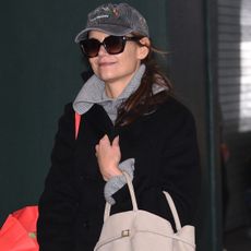 Katie Holmes wears a baseball cap, large sunglasses, gray turtleneck sweater, black coat, and beige bag.