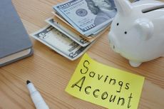 "savings account" written on yellow post-it surrounded by piggy bank, money, and desk supplies