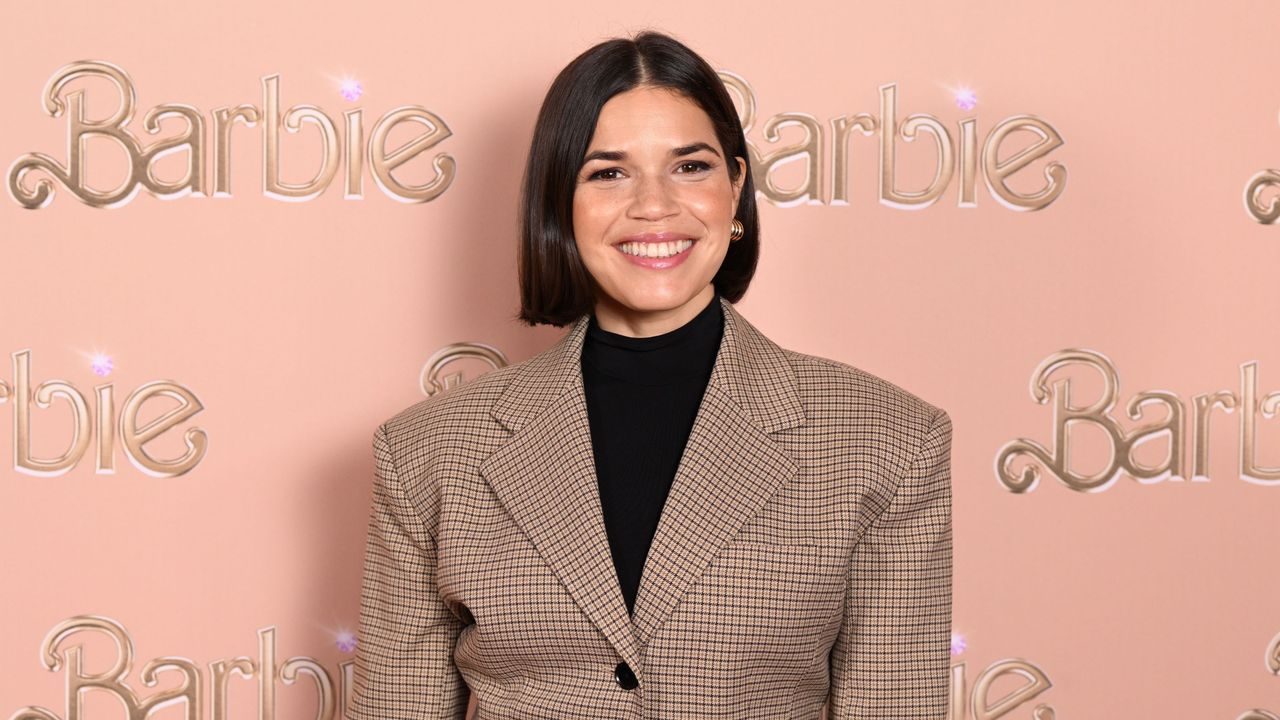 America Ferrera wearing a brown blazer and black turtleneck, standing against a pink background with gold letters that say &#039;Barbie&#039;