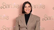 America Ferrera wearing a brown blazer and black turtleneck, standing against a pink background with gold letters that say 'Barbie'