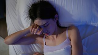 A woman lying in bed with covers pulled over her. She holds the back of one hand up to her face as if just waking up, and her expression looks tired