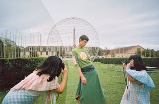 Women photographing a model wearing green with large metal sculpture in background