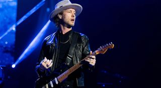 Kenny Wayne Shepherd in wears a wide-brimmed hat and leather jacket onstage and plays a Fender Stratocaster under the stage lights.