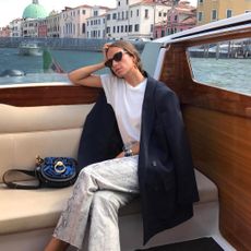 Italian fashion influencer Erika Boldrin sitting on a boat in Venice wearing black cat-eye sunglasses, red earrings, a white t-shirt, oversize black blazer, black patent bag, and snakeskin pants
